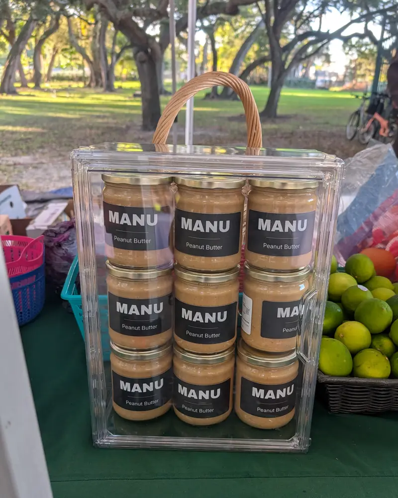 An acrylic case of nine MANU jars on the Urban Oasis Project market table under oak trees.
